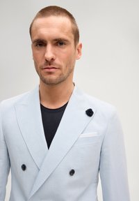 Homme aux cheveux courts portant un blazer croisé bleu clair sur une chemise noire, debout devant un fond gris uni.