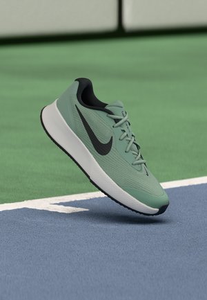 Chaussure de course Nike verte avec un swoosh noir sur un terrain de sport bleu et vert, montrée en plein air.