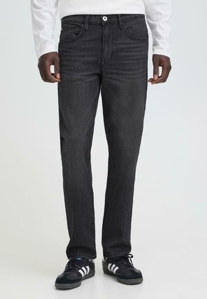 Man draagt zwarte slim-fit jeans, wit langarmshirt en zwarte sneakers met witte strepen en staat tegen een effen achtergrond.