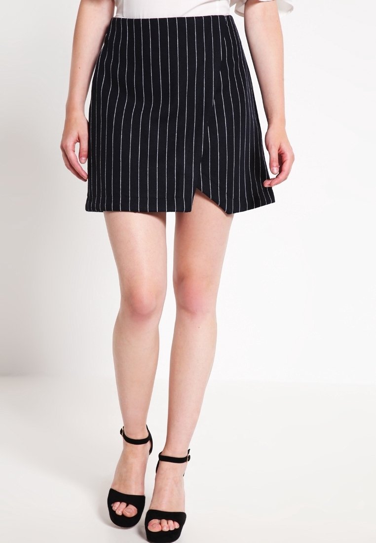 Femme portant une mini-jupe noire à fines rayures avec une fente à l'avant et des sandales à talons hauts noires avec bride à la cheville, debout sur un sol blanc.