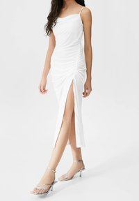 Bílá bezramínková šaty se sklady, bočním rozparkem a hladkým materiálem. Doplněno o stříbrné sandály na vysokém podpatku s nízkým podpatkem.