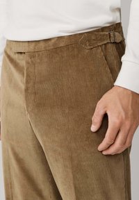 Bruine corduroybroek met verticale ribbels, een vlakke tailleband met een detailsluiting aan de zijkant en een gladde textuur. Hand rustend op de stof.