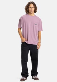 T-shirt a manica corta dal taglio loose-fit in cotone viola chiaro con taschino sul petto; indossato con jeans neri e sneaker marroni.