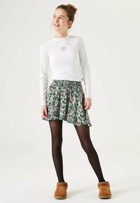 Weißes langärmliges Oberteil mit geribbter Textur und kleinem Logo, kombiniert mit einem bunten Rock im Blumenmuster und schwarzen Strumpfhosen. Braune Hausschuhe mit Fellfutter.