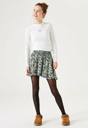 Witte langemouwen top met ribbelstructuur en klein logo, gecombineerd met een kleurrijke rok met bloemenpatroon en zwarte panty’s. Bruine pantoffels met een voering van bont.
