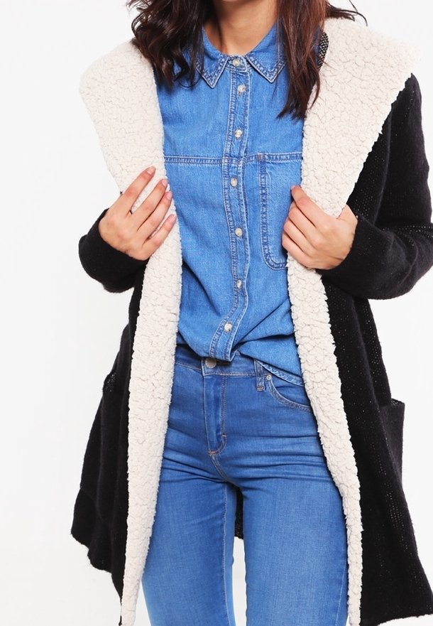 Personne portant une chemise en denim bleu et un jean, tenant un manteau noir ouvert avec une doublure en polaire blanche.