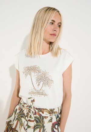 Blonde Frau trägt ein weißes ärmelloses Top mit goldenem Palmenmotiv und gemusterte tropische Shorts mit gebundenem Gürtel.