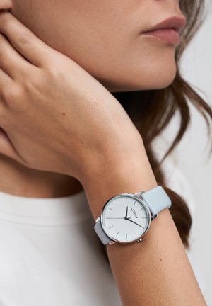 Hellblauer Silikonarmbanduhr mit rundem silbernem Gehäuse, weißem Zifferblatt und minimalistischen schwarzen Stundenmarkierungen. Am Handgelenk getragen.