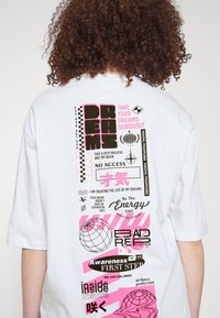 Bílý oversized tričko s grafickým textem a vzory v černé a růžové barvě. Design obsahuje fráze o snech a energii, s ikonami globů.