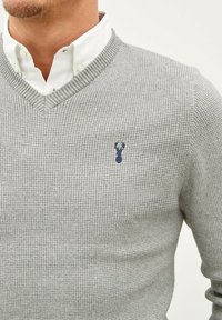 Pull en tricot texturé gris avec un col en V et un petit logo navy sur la poitrine gauche, superposé sur une chemise blanche à col.