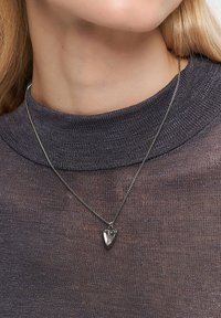 Metallisk halskæde med et hjerteformet vedhæng, sølvfarvet kæde og struktureret grå sweater. Vedhænget har en klar midte med en metallisk ramme.