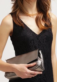 Sac à main clutche en argent métallique avec une texture lisse et un rabat repliable, tenu dans la main. Robe noire à motif en dentelle visible en arrière-plan.