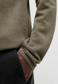 Texturerad olivgrön tröjärmsärm med en åtsittande mudd, som avslöjar en del av en hand dold i en svart byxficka.