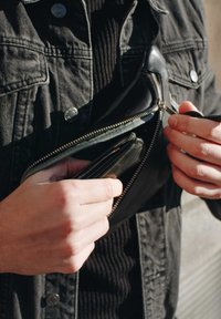 Black leather wallet being retrieved from a zippered pocket of a dark denim jacket, showcasing the wallet's smooth texture and compact shape.