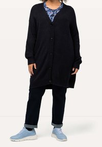 Larga chaqueta de punto de color azul marino oscuro con escote en V, botones en la parte delantera, puños acanalados y aberturas laterales, combinada con una blusa floral azul y zapatillas ligeras.