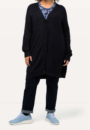 Cardigan long bleu marine foncé avec un décolleté en V, une fermeture à boutons, des poignets côtelés et des fentes latérales, porté avec un haut bleu à motifs floraux et des baskets claires.