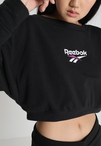 Sudadera recortada negra hecha de material suave, con un logo de Reebok en blanco y morado en el frente. Mangas de corte suelto, diseño minimalista.
