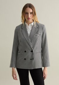 Blazer a cuadros en blanco y negro con diseño de doble botonadura, solapa con muesca y dos bolsillos frontales. Tela suave con silueta estructurada.