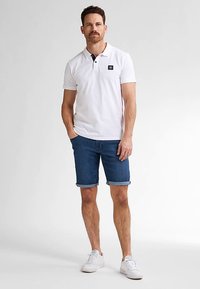 Valkoinen poolopaita, jossa musta kaulus ja lyhyet hihat. Yhdistetty rullattuihin farkkushortseihin. Malli pukeutunut valkoisiin lenkkareihin, seisoo harmaata taustaa vasten.