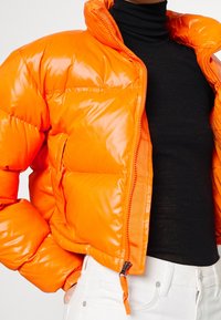 Błyszcząca pomarańczowa kurtka puchowa, rozpięta na czarnym golfie i białych spodniach, z jedną ręką w kieszeni, skrócona do połowy torsu i szyi.