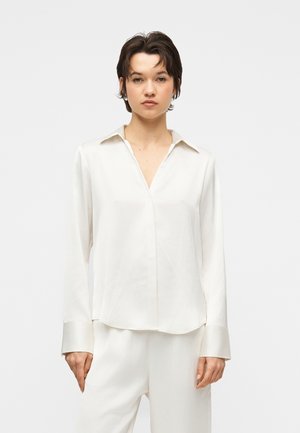 Kvinde med kort mørkt hår iført en elfenbensfarvet langærmet bluse med krave og matchende bukser, stående foran en ensartet lys baggrund.