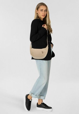 Bolso beige para el hombro con textura lisa, forma curva y logotipo en relieve. Combinado con vaqueros enrollados de color azul claro y zapatillas negras.