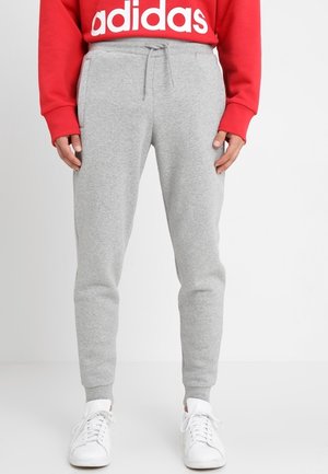 Osoba w czerwonej bluzie Adidas, jasnoszarych spodniach dresowych ze sznurkiem oraz białych trampkach, stojąca na tle jednolitego jasnego tła.