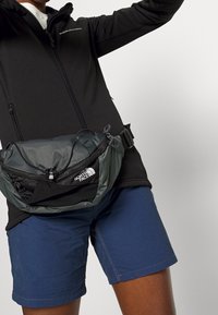 Chaqueta negra con cremallera, pantalones cortos azules y una riñonera gris con un acento negro. La riñonera tiene un cierre con cremallera y una correa ajustable.