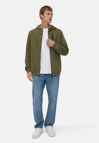 Veste à capuche zippée olive avec une capuche, poches avant et détails de fermeture éclair noirs, portée par-dessus un t-shirt blanc et un jean bleu, avec des chaussures blanches.