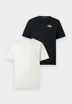 Mann mit schwarzen Sonnenbrillen, schlichtem weißen T-Shirt, schwarzen weit geschnittenen Hosen und grau-weißen Turnschuhen steht vor weißem Hintergrund.