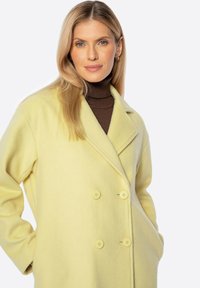 Abrigo de mezcla de lana amarillo, de doble botonadura, con grandes botones, solapas de muesca y textura suave. Llevado sobre un suéter marrón de cuello alto.