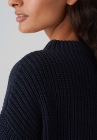 Pull en maille bleu marine foncé, doté d'une texture côtelée et d'un col montant structuré, montrant une vue latérale de l'épaule et du col.