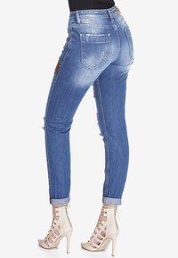 Jeans de denim azul desgastado con un ajuste slim, ligeras desgastes y bordado floral en el muslo. Los dobladillos enrollados revelan tiras en los tobillos de tacones beige.