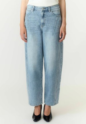 Femme portant un jean bleu clair taille haute à jambes larges avec des détails de couture à l'avant et des talons noirs à bout pointu, debout devant un fond uni.