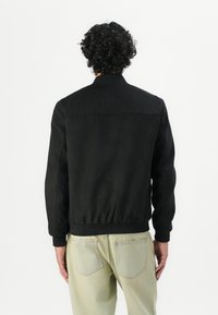 Svart bomberjakke med jevn tekstur, elastiske mansjetter og liv, samt en enkel kragedesign. Båret med lyse jeans.