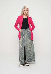 Cardigan rosa a maglia sopra una maglietta nera, abbinato a una lunga gonna in denim grigia con stampa pitone, caratterizzata da strappi e uno spacco anteriore. Sneakers nere.