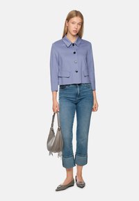 Light purple cropped jacket with a collar, black buttons, two front pockets; paired with light blue jeans, grey handbag, and grey flats.