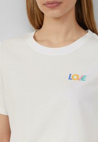 Witte katoenen t-shirt met een ronde halslijn, waarop het woord "LOVE" staat in meerkleurige letters: blauw, oranje, geel en groen.