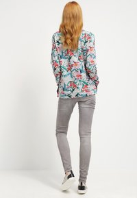 Camisa de manga larga floral con base gris, que presenta vibrantes patrones en rosa y verde; combinada con jeans ajustados gris claro y zapatos slip-on.
