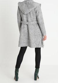 Grå ullkappa med huva, bältad i midjan, med en strukturerad yta, sidofickor och kombinerad med svarta jeans och gröna klackstövlar.