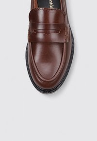 Mocasines de cuero marrón con punta redondeada, tira decorativa, detalles de costura y una suela de goma negra texturizada.