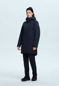Parka impermeabile blu marina con cappuccio, tasche con zip e patch con logo a contrasto. Indossato sopra pantaloni neri e scarponi da escursionismo neri.