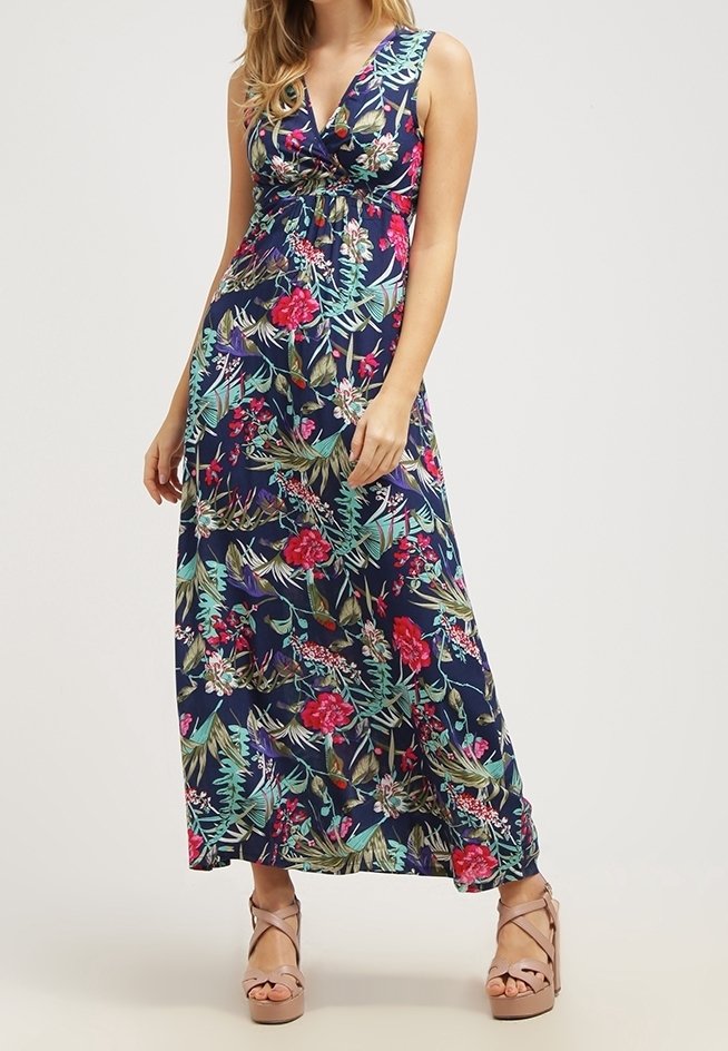 Femme portant une robe maxi sans manches bleu marine avec des motifs floraux colorés et des sandales compensées beige, debout devant un fond uni.