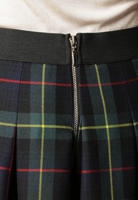 Falda de tartán con una cinturilla elástica negra y un cierre de cremallera plateada. La tela presenta un patrón de colores verde, amarillo, rojo y negro.
