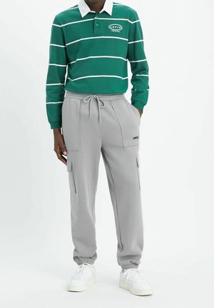 Person trägt ein grün-weiß gestreiftes langärmeliges Polo, hellgraue Cargo-Jogginghosen mit Kordelzug und Levi’s-Logo sowie weiße Sneakers.
