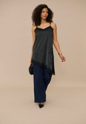 Femme aux cheveux bouclés portant un haut noir sans manches avec bordure en dentelle, un pantalon large foncé, des chaussures noires pointues et tenant un sac à main noir.