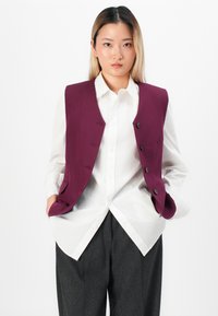 Jeune femme avec des cheveux raides portant une chemise blanche à boutons, un gilet violet à boutons et un pantalon gris foncé, se tenant les mains dans les poches.