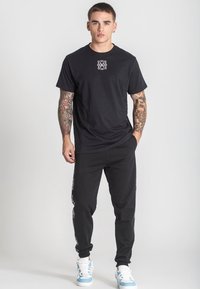 Svart bomulls-t-shirt med en vit grafik på bröstet, kombinerad med svarta joggers som har ett mönstrat sidaccent och vita sneakers.