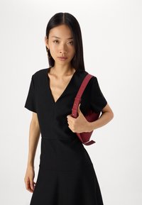 Vestuário preto com mangas curtas e decote em V, apresentando um corpete justo que se alarga na cintura. Segurando uma bolsa vermelha com alça.