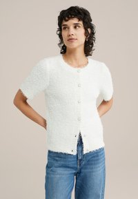 Vit kortärmad kofta i texturerat, fluffigt material med rund halsringning och fem pärlliknande knappar framtill.